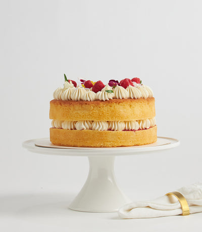 Three-tier raspberry sponge cake with piped white buttercream, fresh raspberries, and edible flowers on ceramic stand