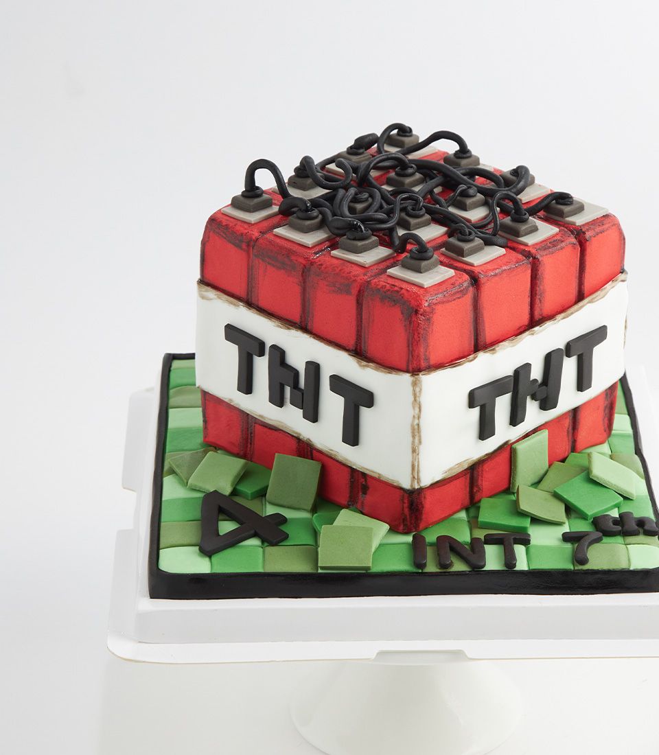 Square red Minecraft TNT birthday cake with white band, black wires, grey detonators, and grass block base for kids'