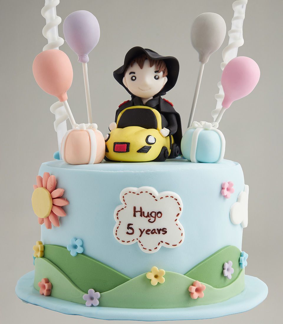 Two-tier fondant birthday cake with toy car driver figurine, pastel balloons, and custom name plaque for children's