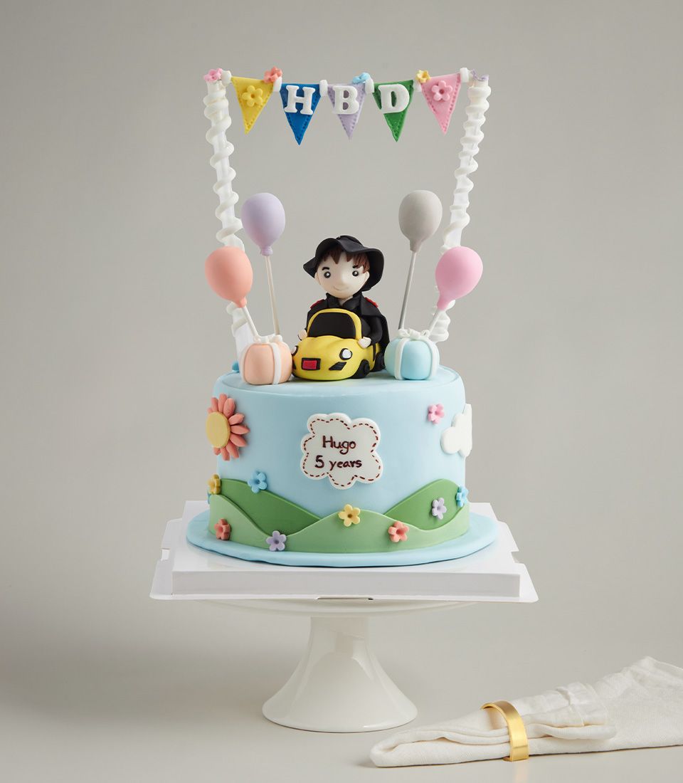 Light blue fondant cake with yellow car and boy figurine, colorful flowers, balloons, and HBD banner for children's birthdays