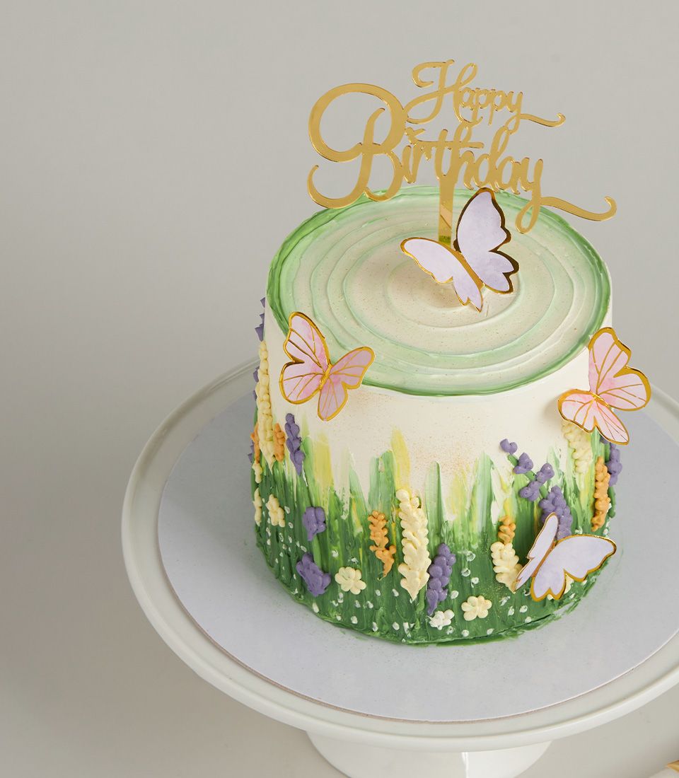 White round birthday cake with green and purple meadow design, pink and gold butterflies, light green frosting top, and gold