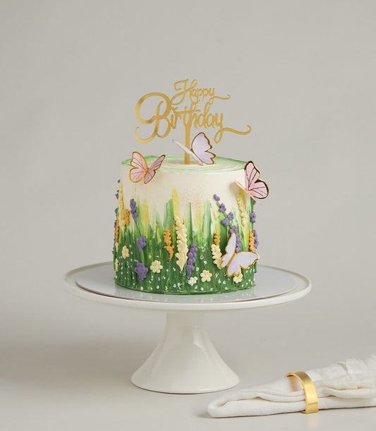 Vibrant green buttercream birthday cake with purple, yellow wildflowers and pink butterfly decorations on ceramic stand
