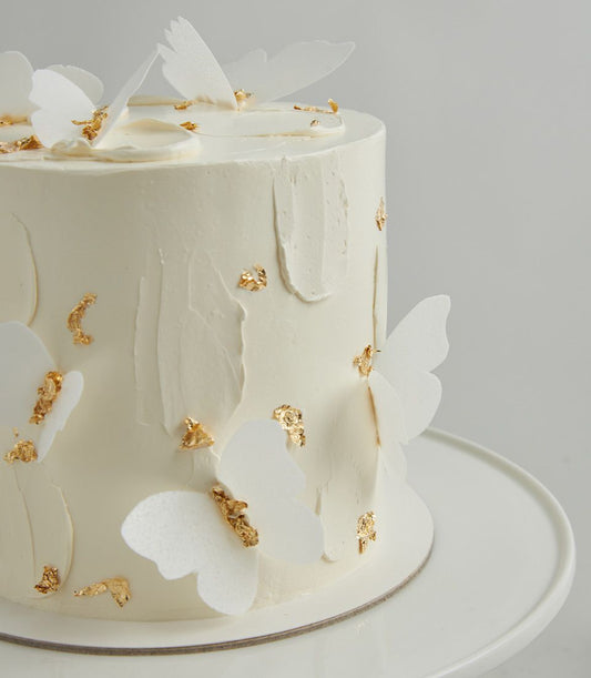 Elegant white layer cake with textured frosting, delicate butterfly decorations, and gold leaf accents for birthdays