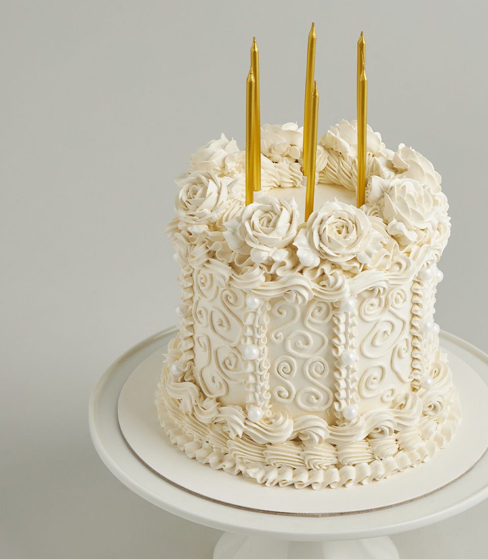 White buttercream cake with piped roses and gold candles on pedestal stand, Charlotte Bakery vintage celebration design