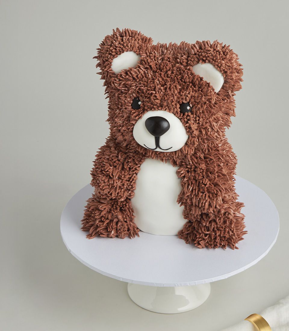 Teddy bear cake with brown fur frosting, white muzzle and inner ears on white cake stand