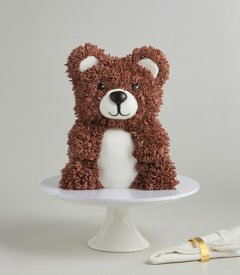 Teddy bear-shaped vanilla cake with brown fur texture, white fondant belly, and black facial features on ceramic stand