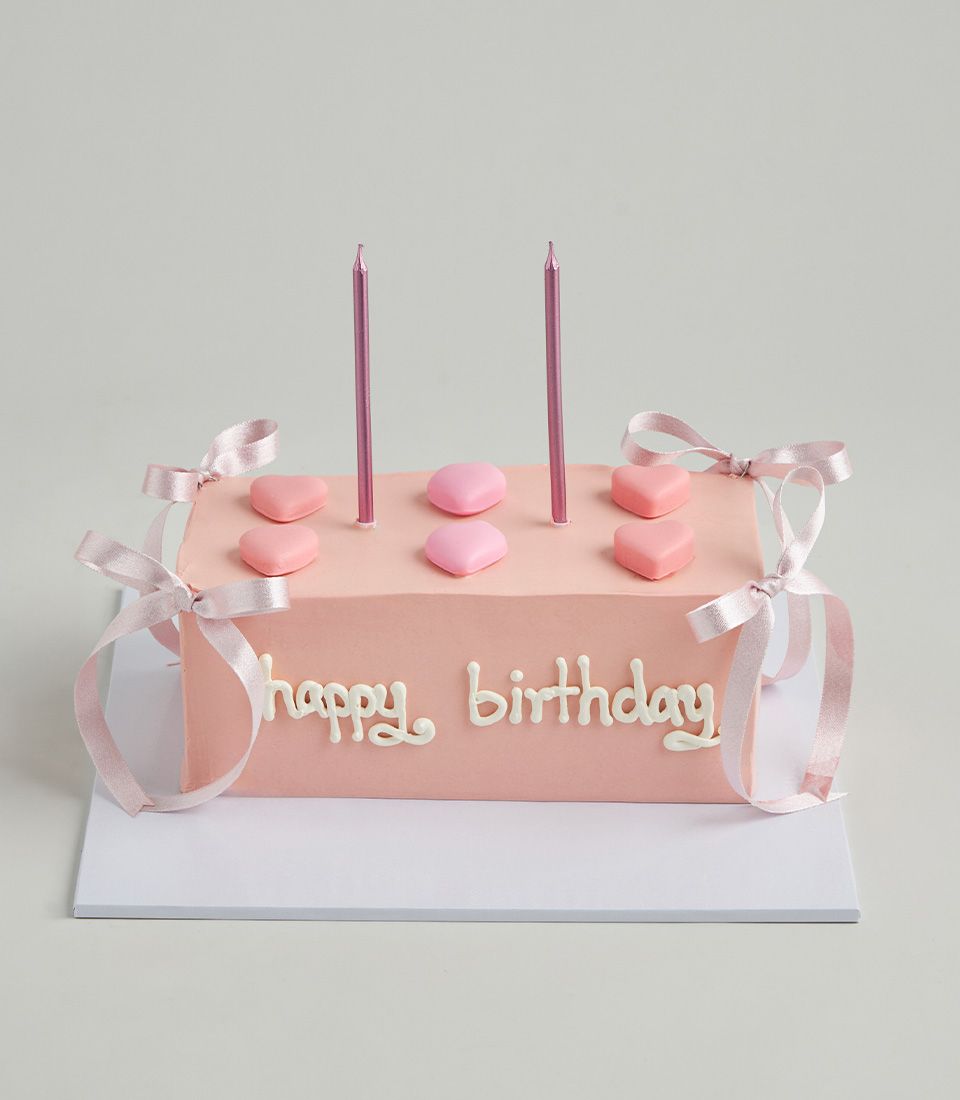 Light pink layer cake with heart candies, ribbon bows, and metallic candles for birthday celebrations