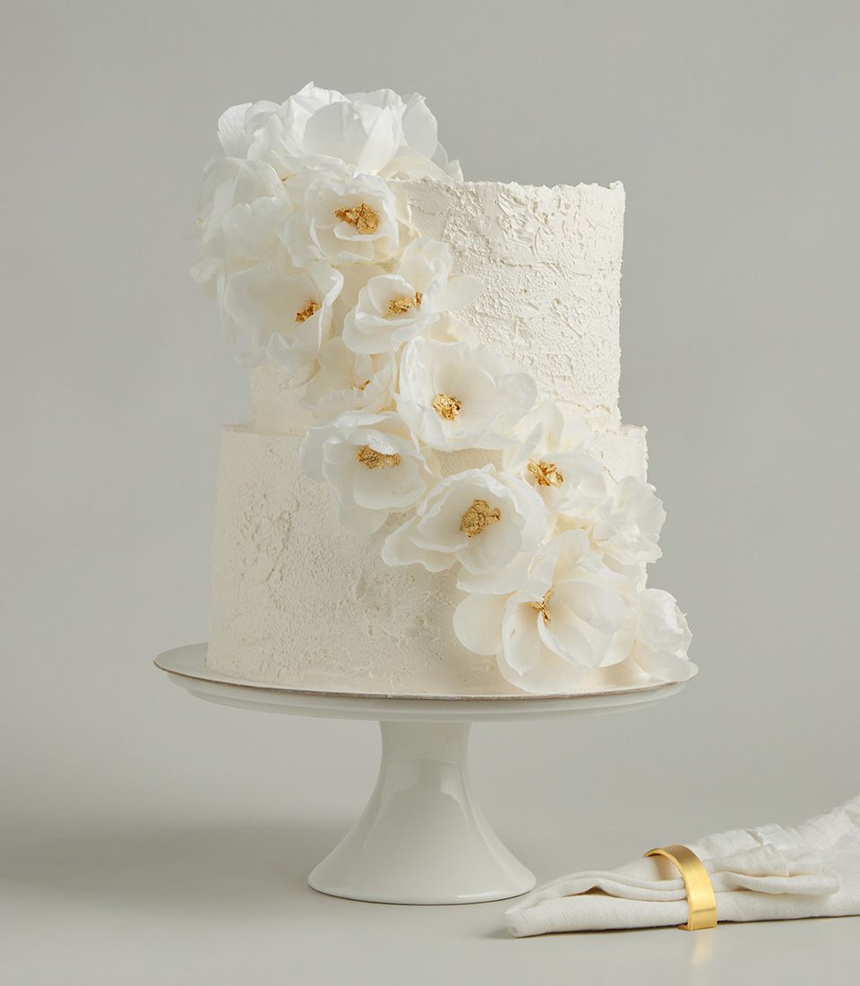 Two-tier white wedding cake with cascading wafer paper flowers and gold leaf detail on rustic buttercream frosting