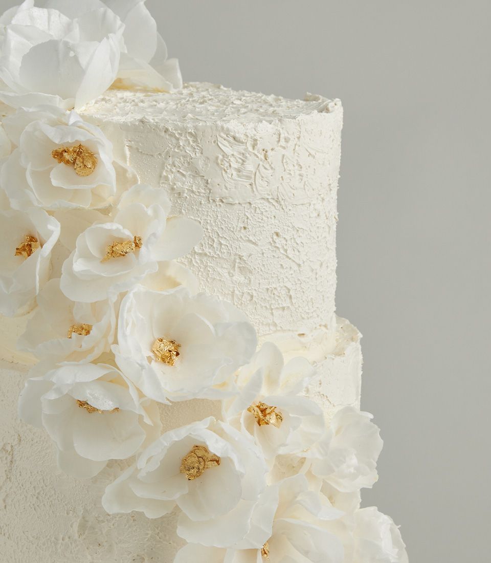 Elegant white two-tier wedding cake with textured buttercream, cascading white edible flowers, and gold leaf accents