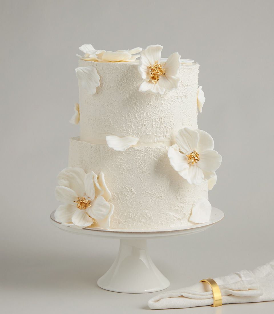 Two-tier white wedding cake with delicate sugar flowers and gold leaf details on ceramic stand
