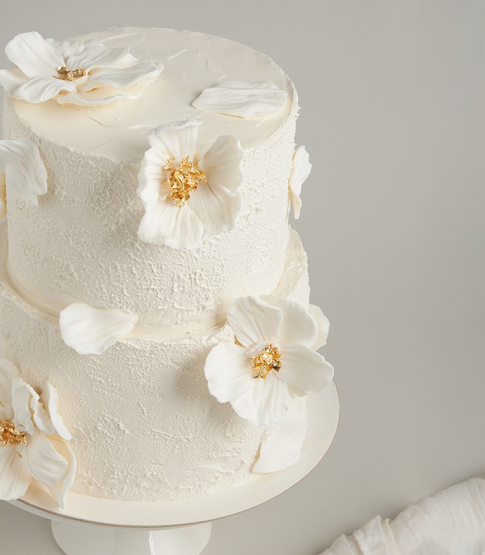 Two-tier white wedding cake with fondant sugar flowers and gold leaf accents on rustic buttercream