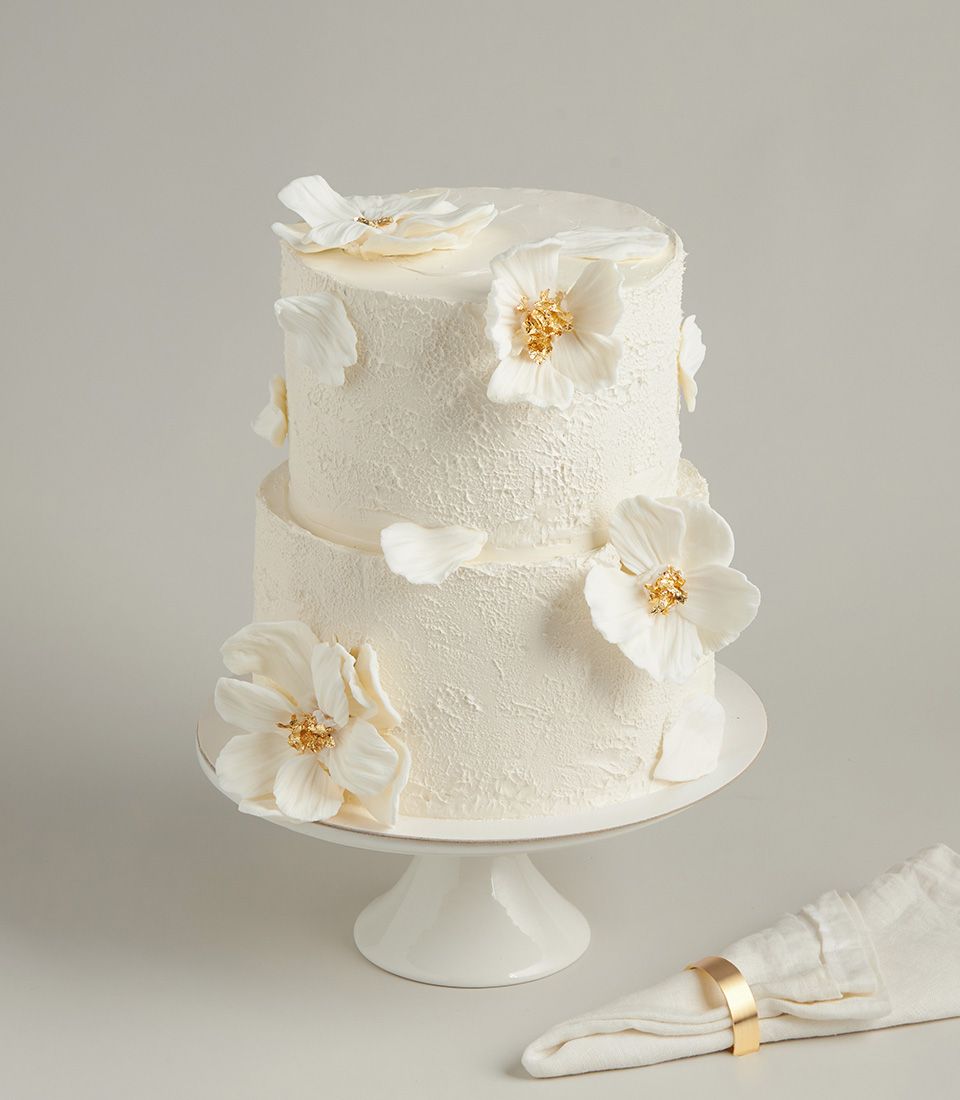 Two-tier white fondant wedding cake with sugar flowers and gold accents on ceramic stand