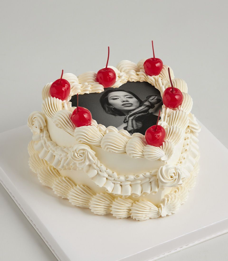 Heart-shaped white layer cake with edible photo, cherry garnish, and ruffled frosting swirls