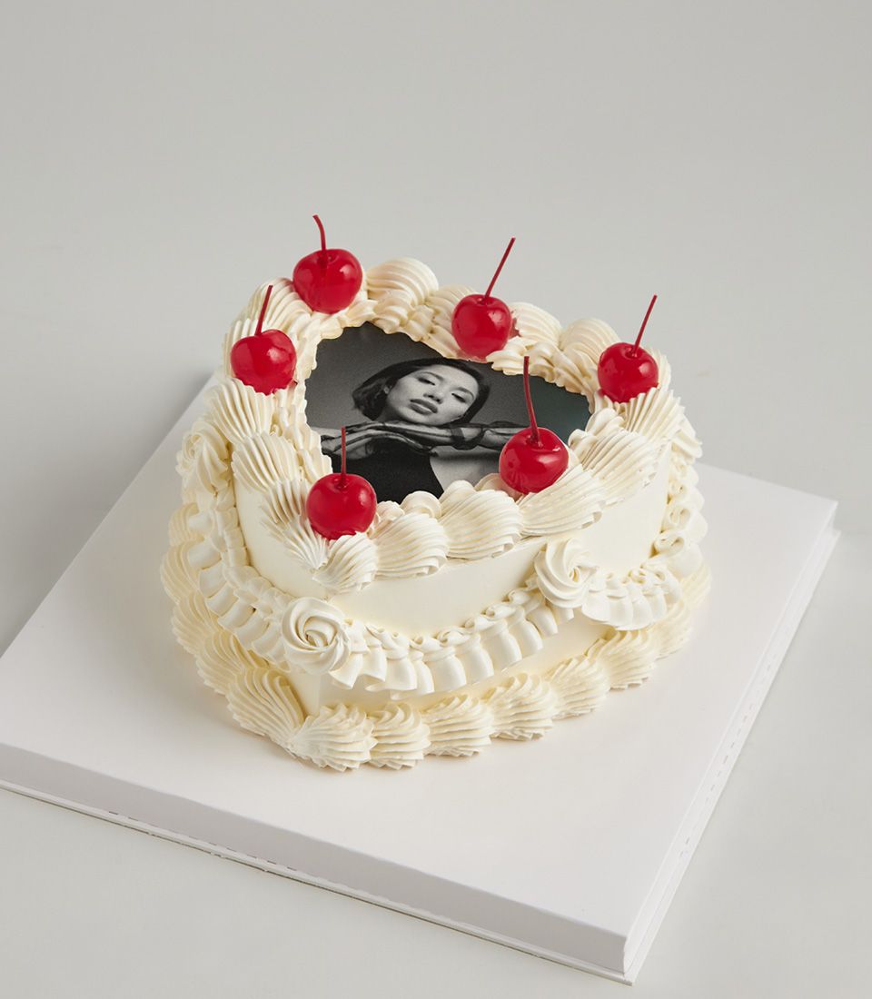 Heart-shaped white cake with edible photo portrait, frosting swirls, and red cherry accents