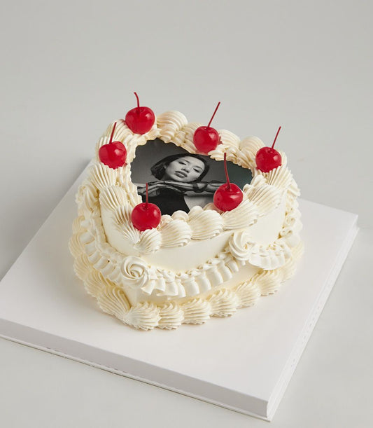 Heart-shaped white cake with edible photo portrait, frosting swirls, and red cherry accents