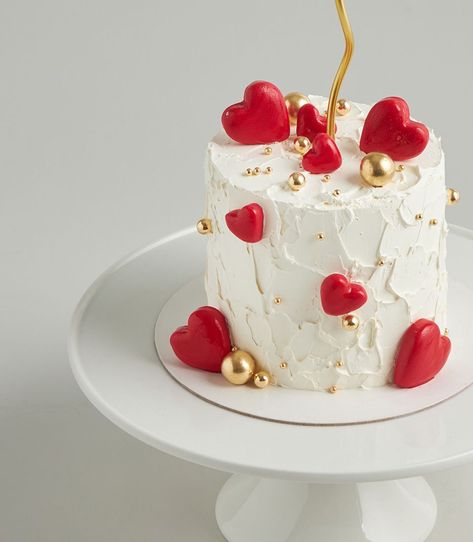 Elegant white celebration cake with red heart candies and gold sprinkles, topped with golden candle accent
