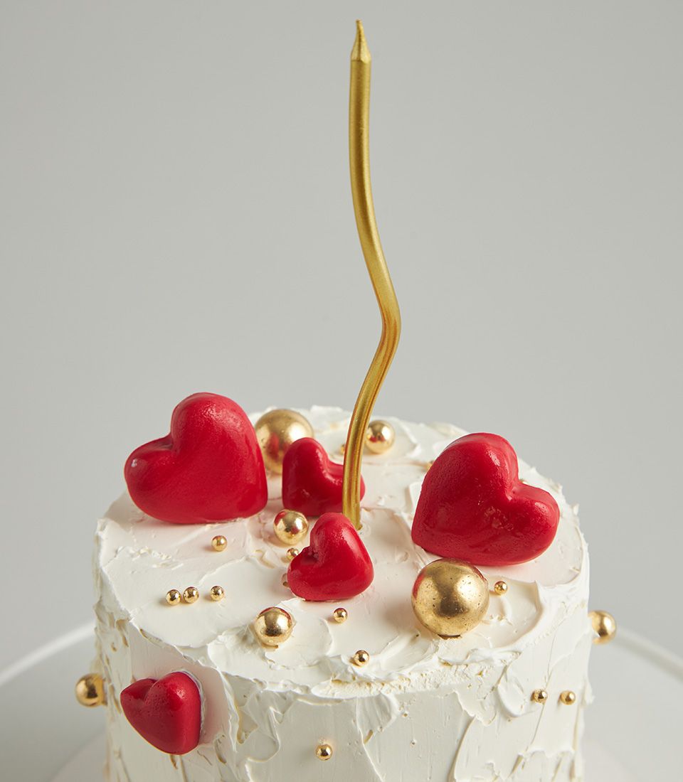 Minimal red heart celebration cake with white frosting, gold spheres, and wavy golden candle on cake stand