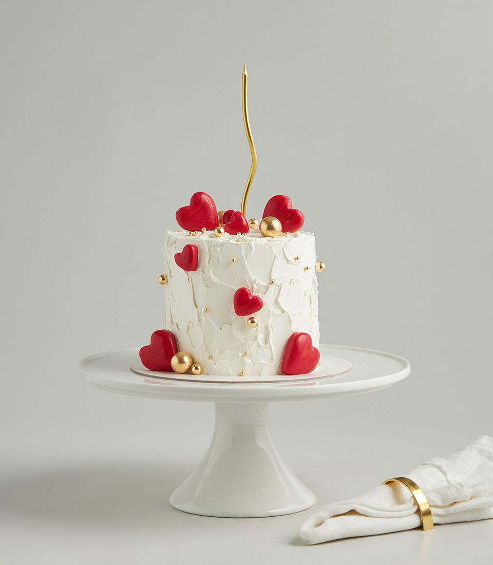 Minimal red heart celebration cake with white frosting, gold pearls, and gold candle on ceramic stand