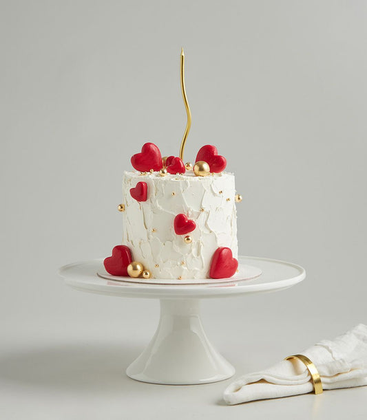 Minimal red heart celebration cake with white frosting, gold pearls, and gold candle on ceramic stand
