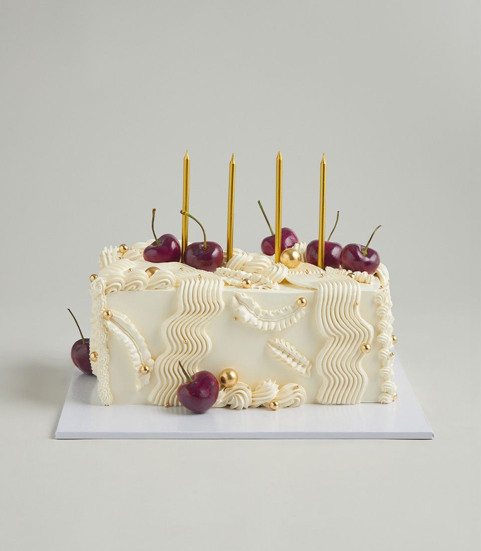 Rectangular white layer cake with cherry and gold piping, fresh cherries, and edible gold accents