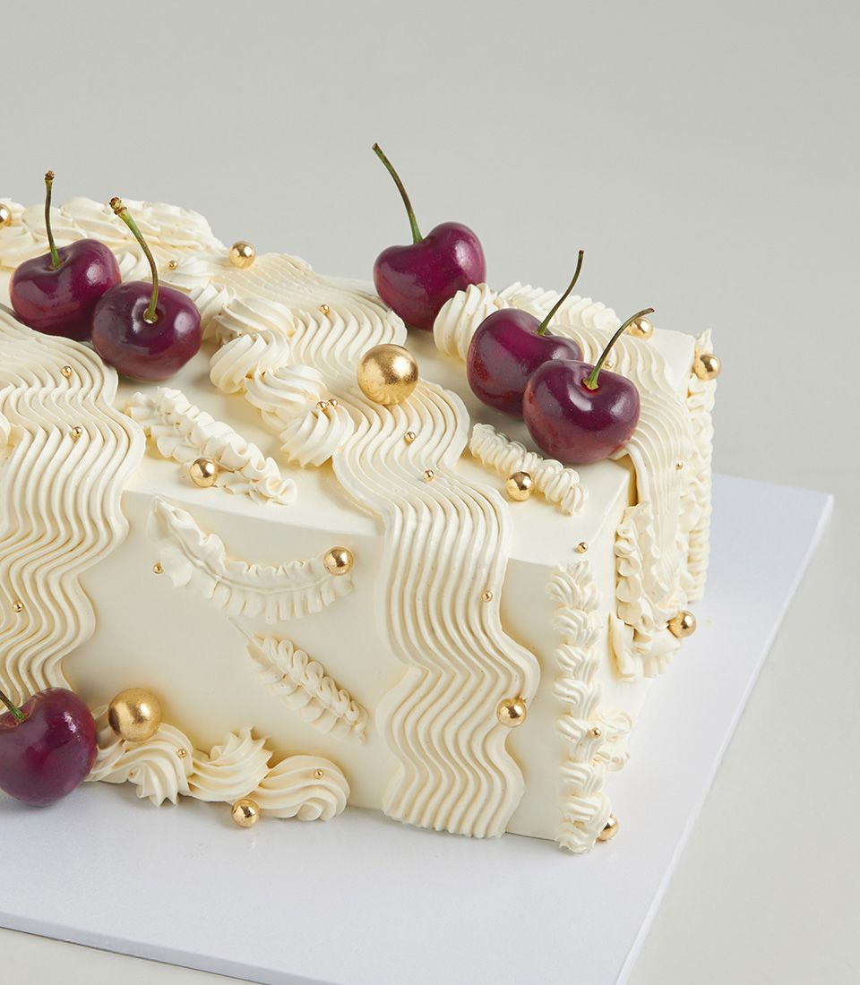 Rectangular cherry cream cake with white frosting, gold pearl piping, and dark red cherries on top