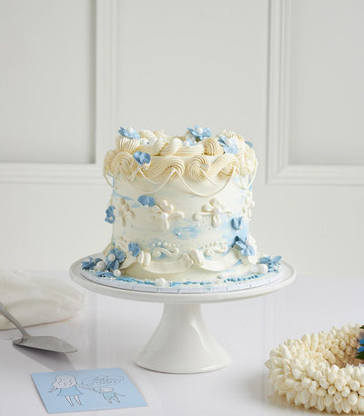 Tiered Mother's Day cake with white frosting, blue watercolor wash, and delicate blue flowers on ceramic stand