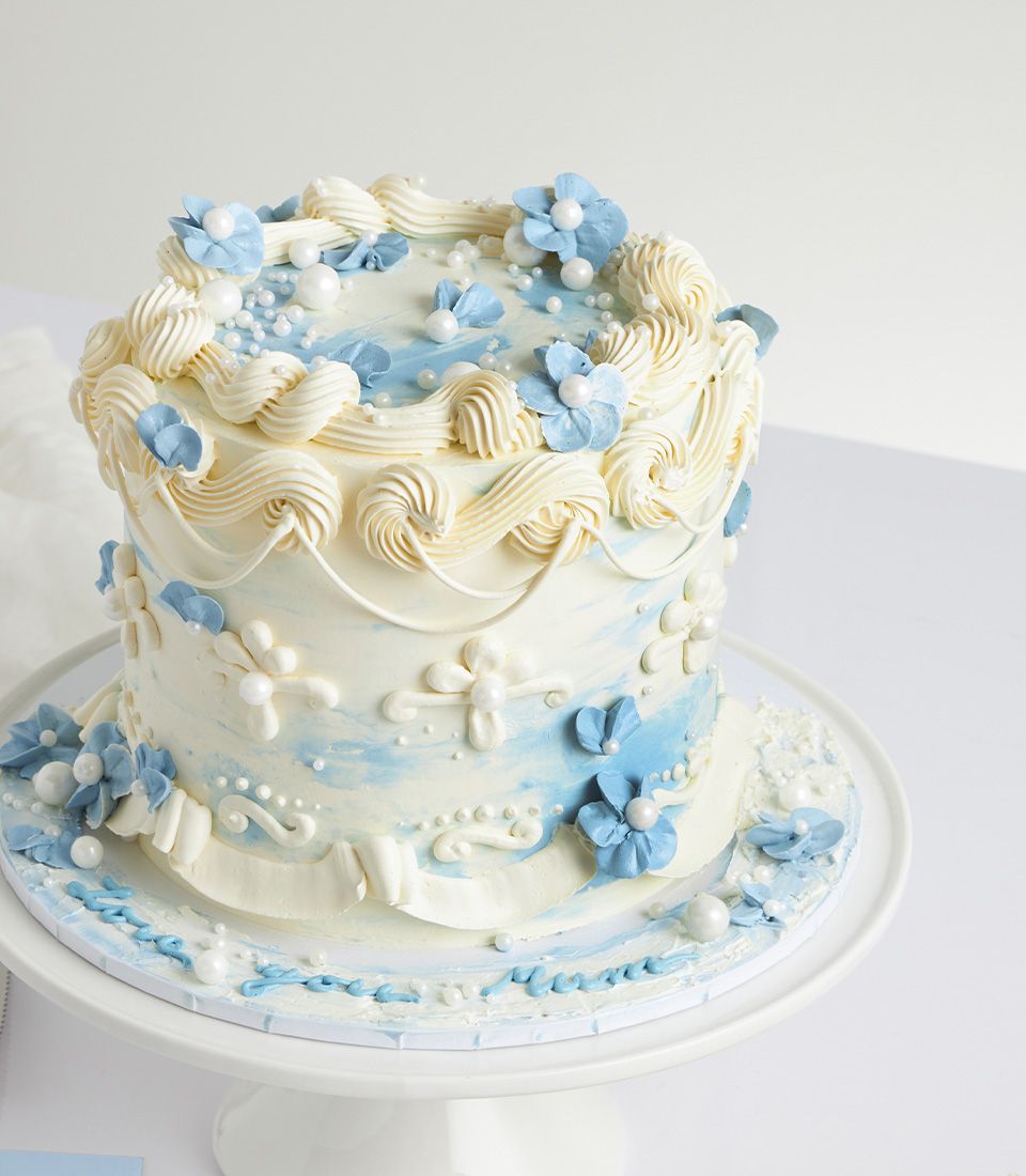 Two-tier Mother's Day cake with white buttercream, light blue watercolor swirls, and pearl-adorned sugar flowers