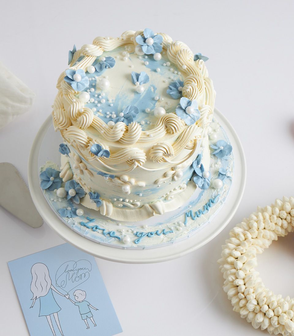 Two-tiered vanilla cake with white frosting, blue floral watercolor details and edible pearls, "Love you Mom" inscription,