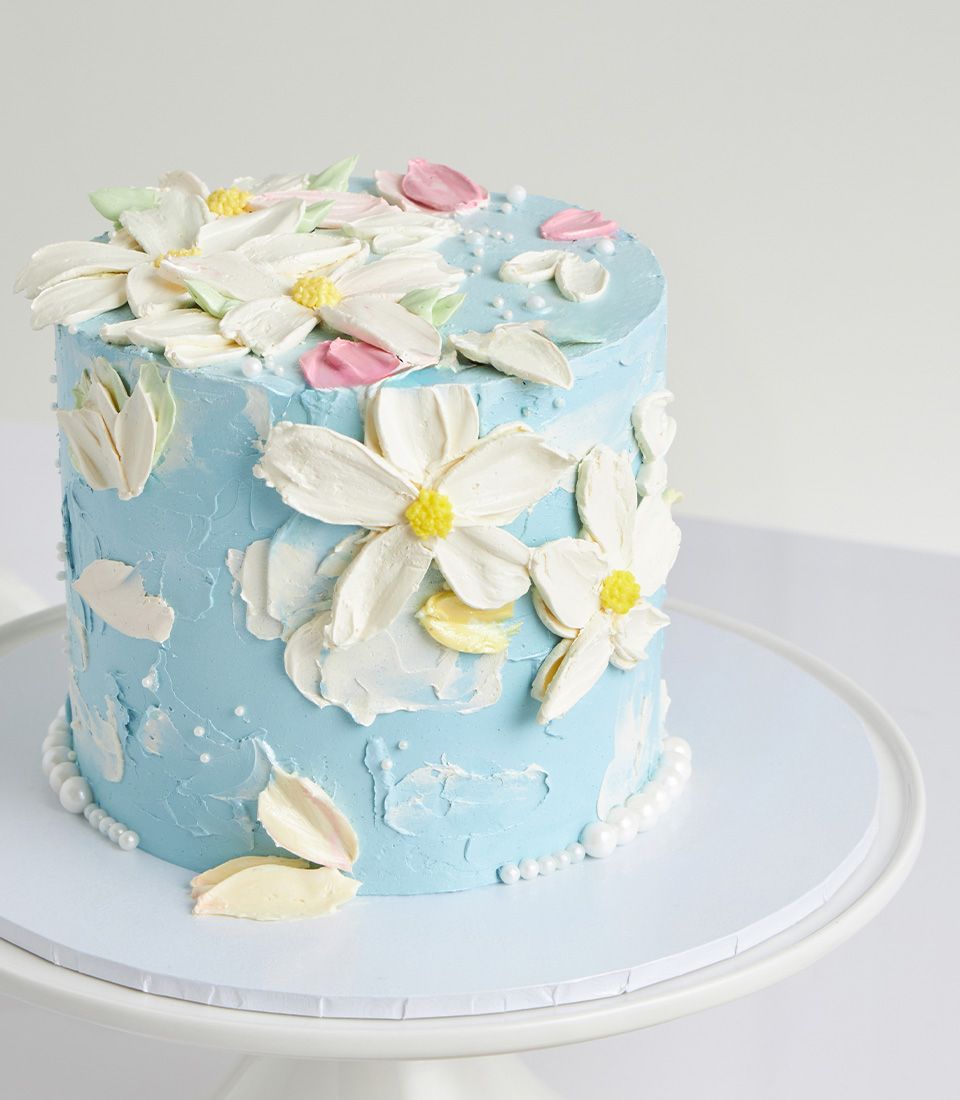 Light blue Mother's Day cake with white, yellow, and pink buttercream flowers on ceramic stand