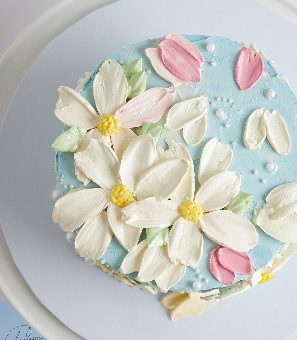 Elegant white and pink buttercream flower cake with yellow centers and edible pearls on light blue frosting