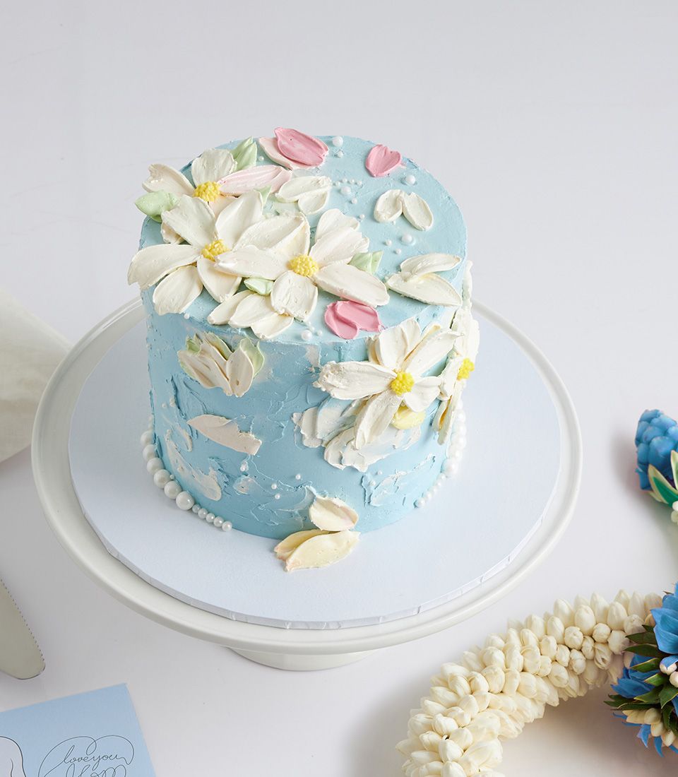 Light blue round cake with white, yellow, and pink floral buttercream, jasmine garland, and Mother's Day card on white stand