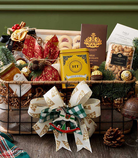 Festive dark metal mesh gift basket with gourmet cookies, mango jam, herbal tea, chocolate, and cashews, decorated with pine