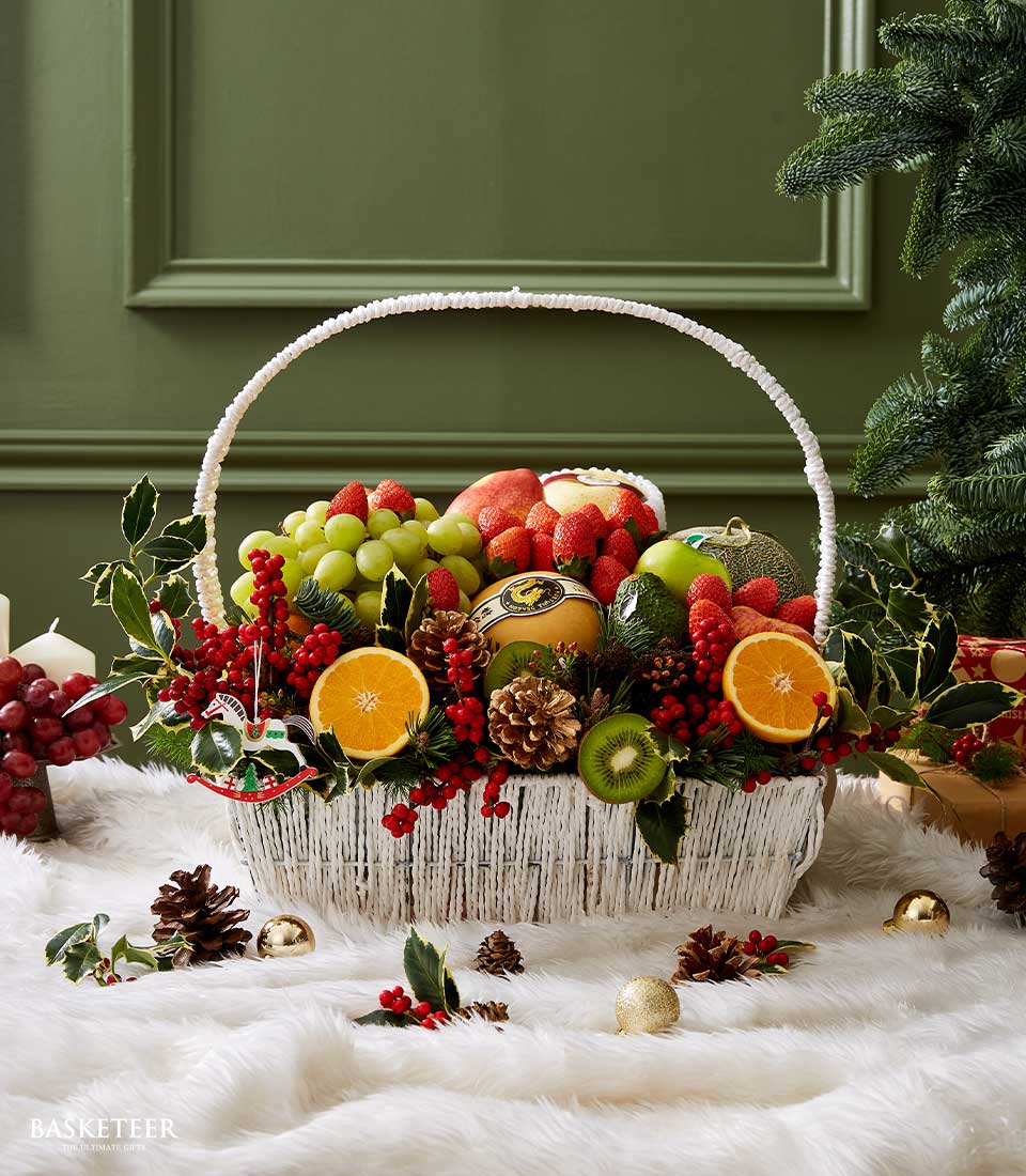 White woven basket overflowing with fresh apples, strawberries, grapes, and pears, accented with holly and pinecones for