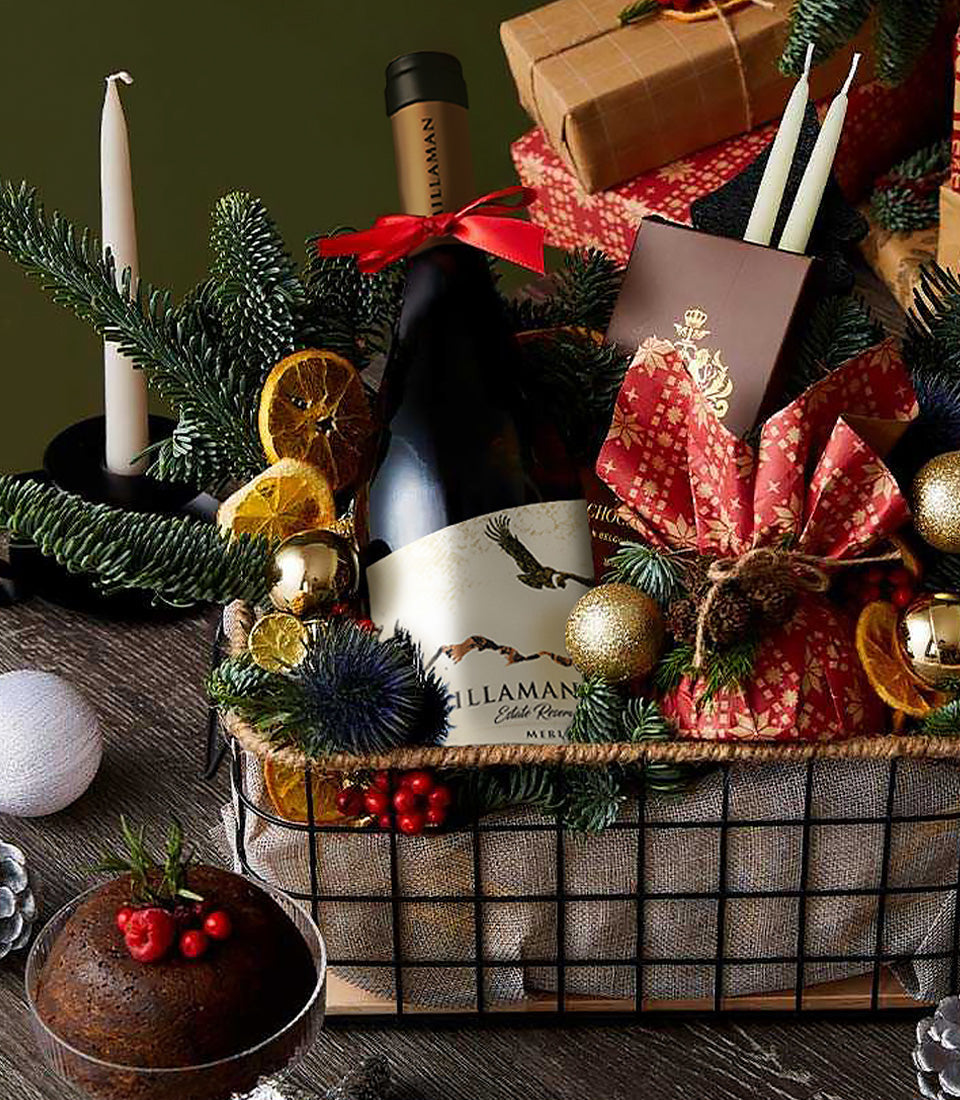 Merlot wine festive basket with red wine, burgundy linens, pine, dried citrus, gold baubles, and holiday dessert