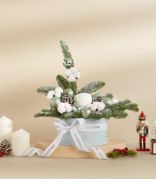 Frosted holiday arrangement with fir branches, white cotton, silver baubles, and pinecones in light blue box with white