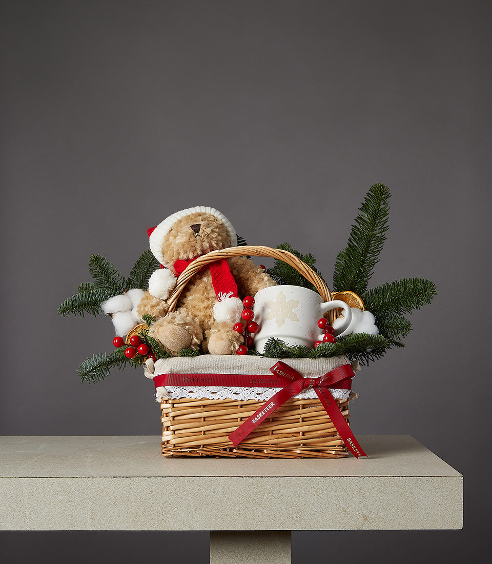 Festive wicker gift basket with light brown teddy bear in Santa hat, ceramic snowflake mug, and holiday greenery