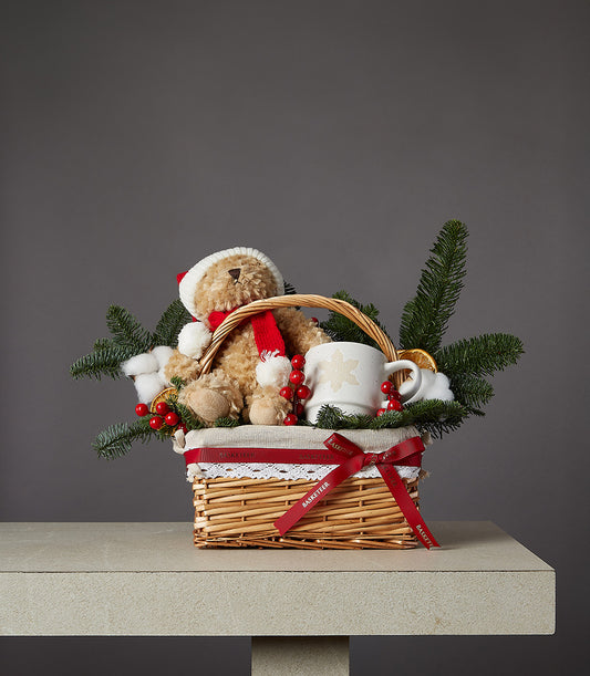 Festive wicker gift basket with light brown teddy bear in Santa hat, ceramic snowflake mug, and holiday greenery