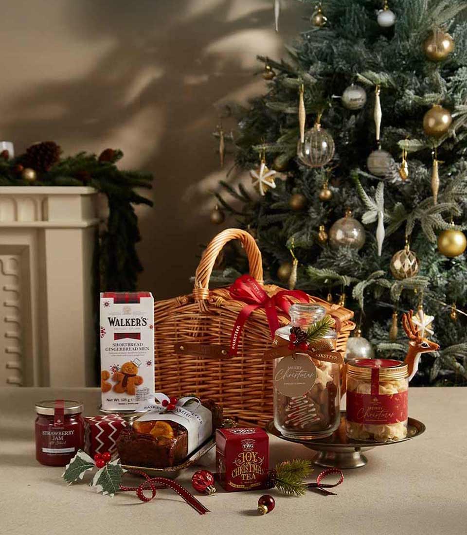 Woven wicker Christmas hamper with red ribbons, shortbread, jam, fruit cake, and festive treats on holiday mantel display