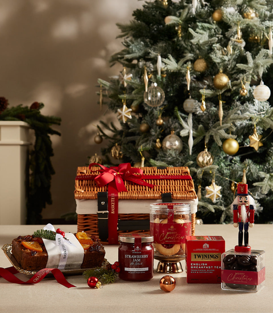 Festive Christmas hamper with fruit cake, jam, tea, cookies, and chocolates in wicker basket with red ribbon