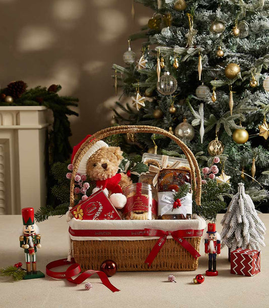 Festive Christmas hamper with teddy bear, Lindor chocolates, cookies, and fruitcake in woven basket with red ribbon
