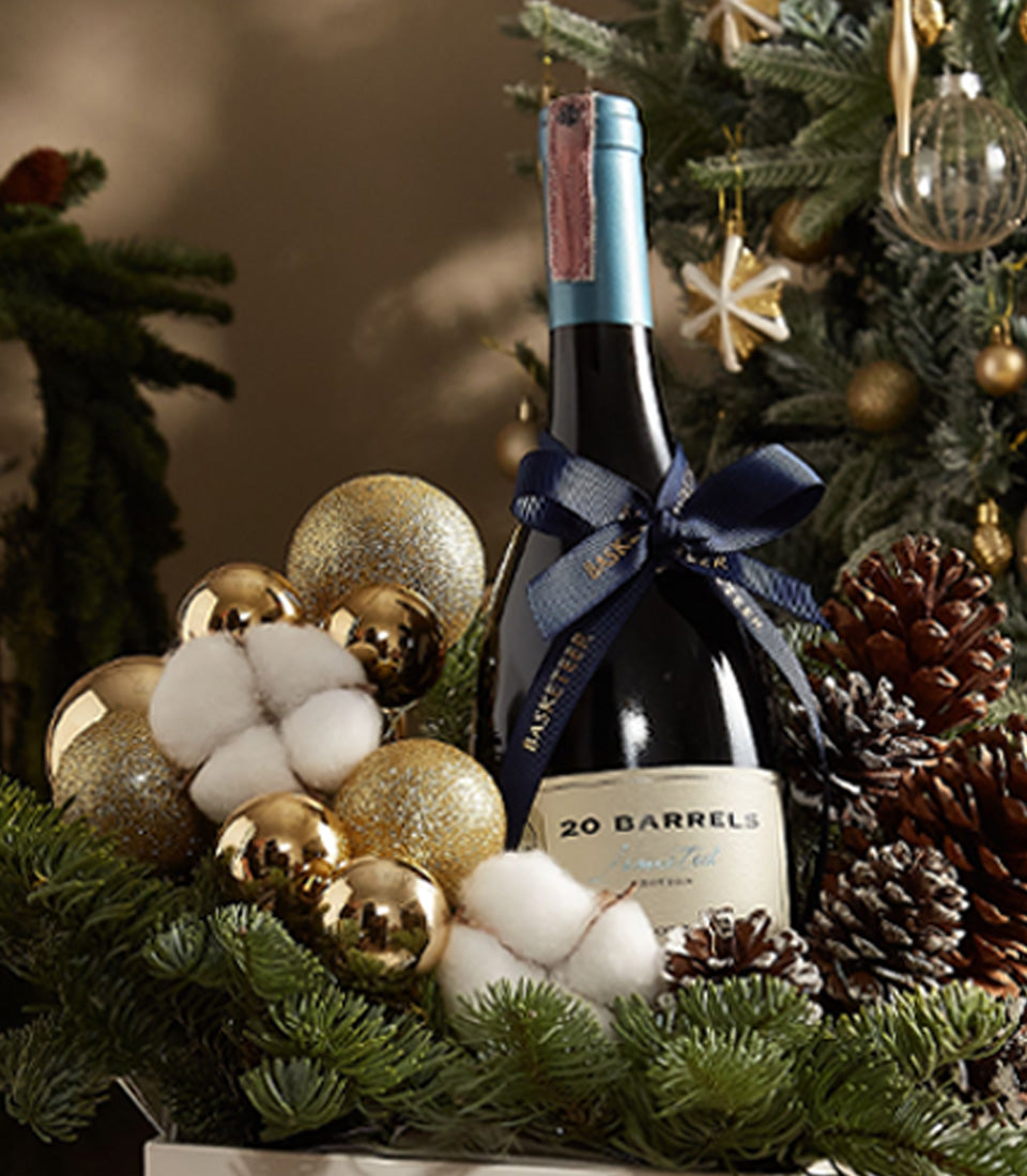 Red wine holiday gift set with gold ornaments, pine branches, and festive ribbon centerpiece