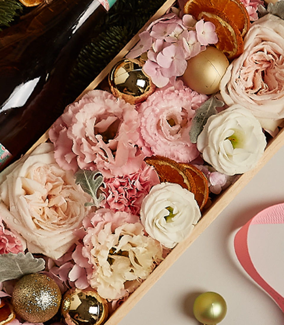 Rosy floral gift box with pink roses, ranunculus, hydrangeas, champagne bottles, and gold Christmas ornaments