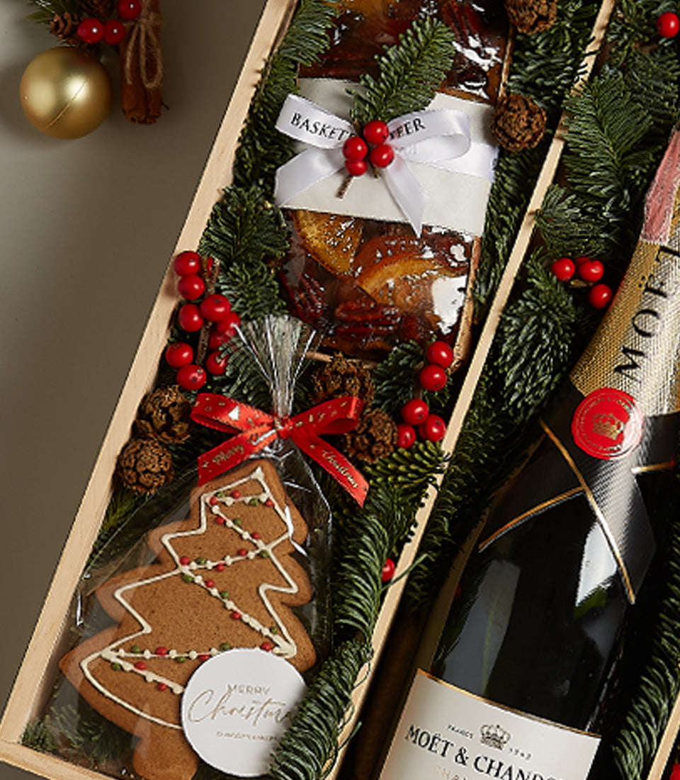 Elegant wooden gift box with champagne, fruitcake, and gingerbread cookie surrounded by festive greenery and red berries