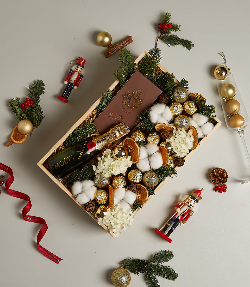 Luxury wooden gift box with Moët champagne, dark chocolate, and gold ornaments nestled in evergreen for Christmas