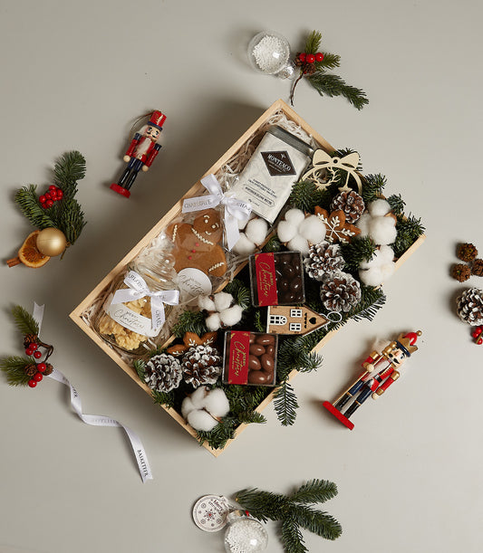 Wooden Christmas gift box with gingerbread cookies, chocolate treats, and festive pine branches on light grey background