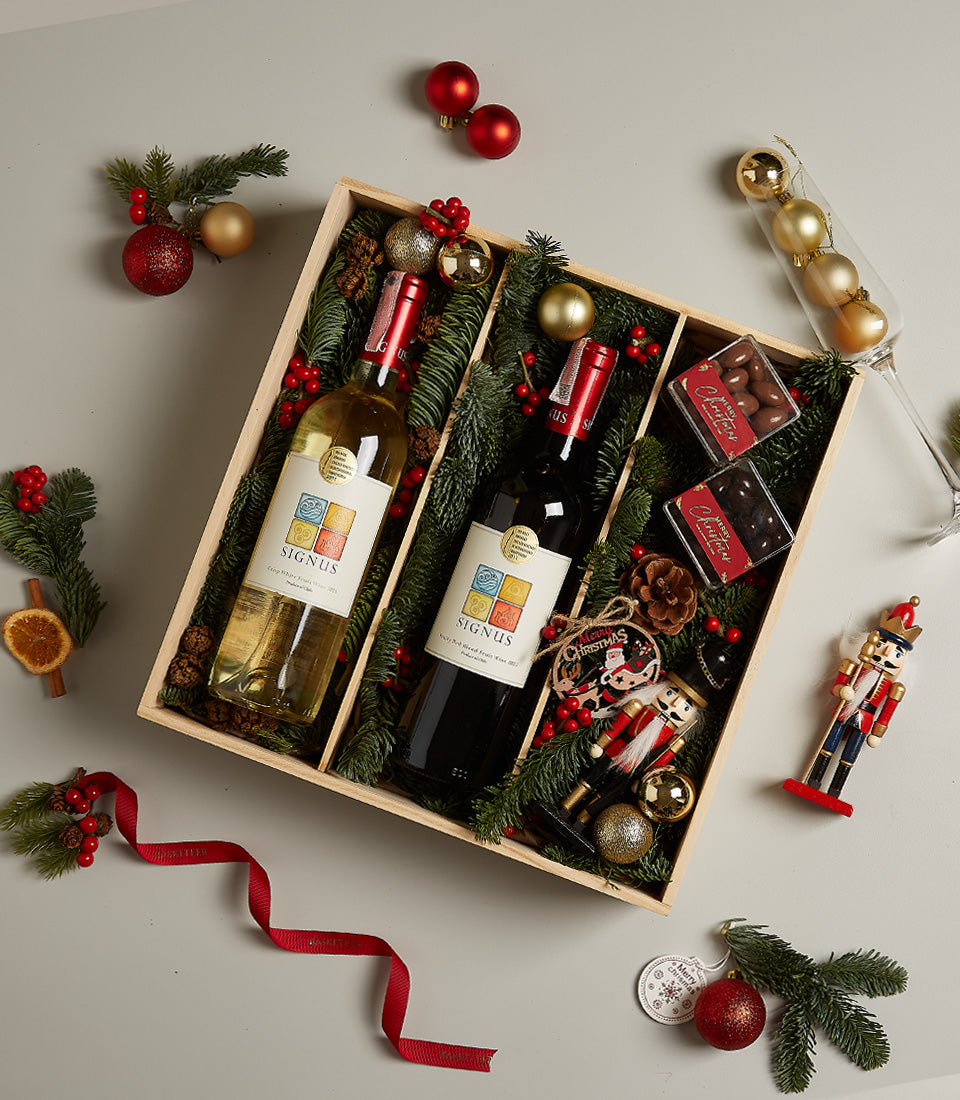 Wooden gift box with SIGNUS red and white wine, chocolate truffles, and festive Christmas ornaments surrounded by pine