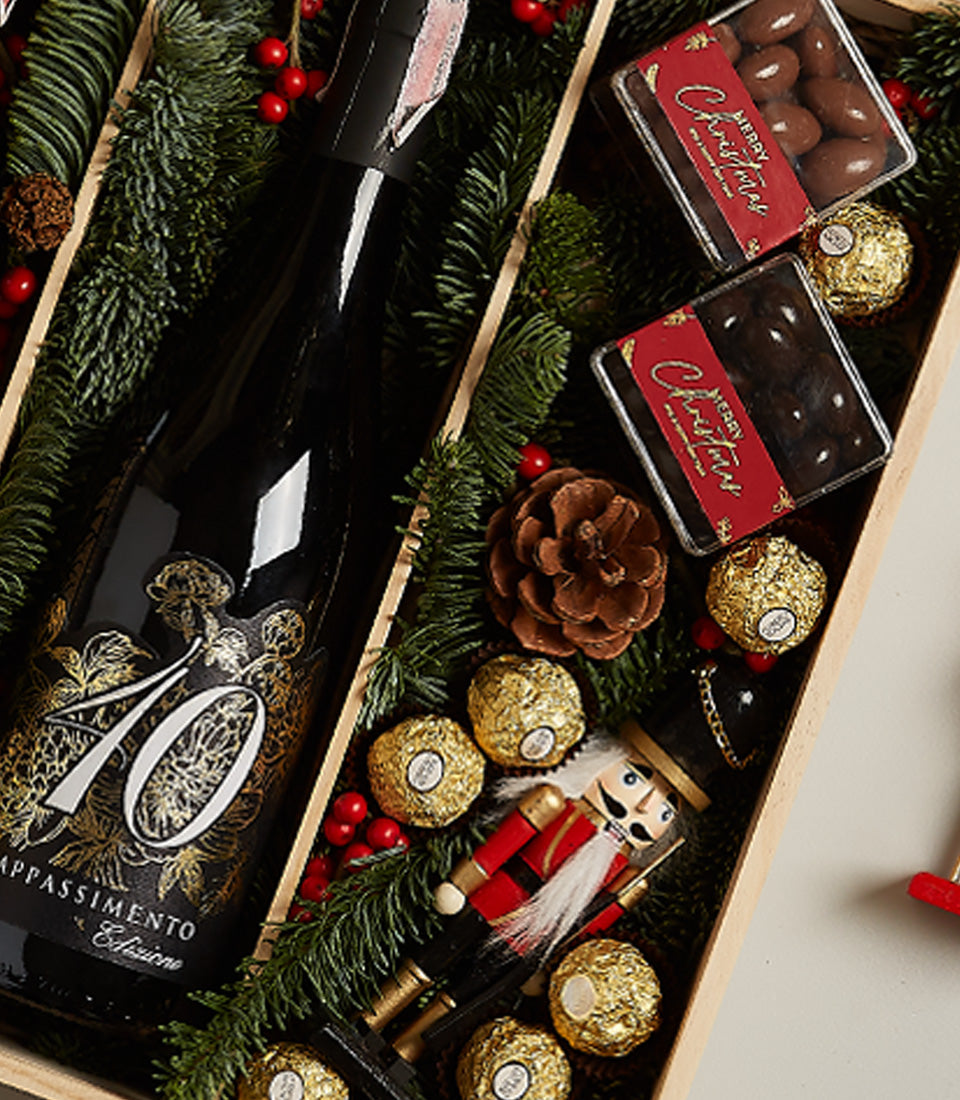 Premium red wine and chocolate holiday gift set with festive green branches, gold foiled chocolates, and wooden nutcracker