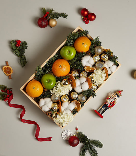 Festive wooden gift box with fresh apples, oranges, Ferrero Rocher chocolates, and evergreen sprigs for Christmas