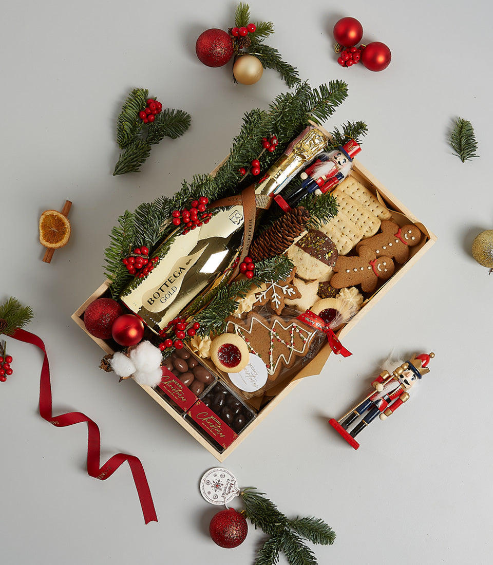 Wooden crate with gold Bottega champagne, gingerbread cookies, Christmas chocolates, and festive pine branches with red