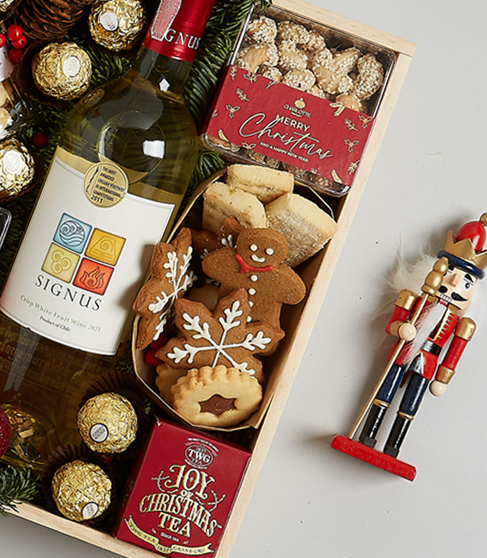 Festive white wine gift hamper with gingerbread cookies, nuts, chocolates, and holiday tea on evergreen base